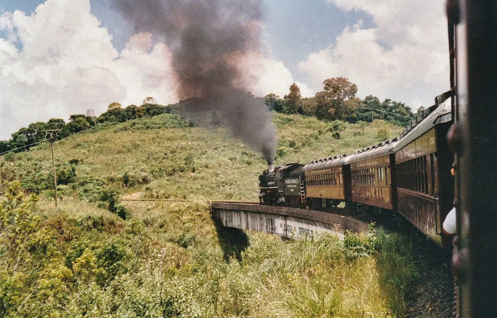 Locomotiva da Maria Fumaça soltando fumaça e puxando os vagões entre morros verdes